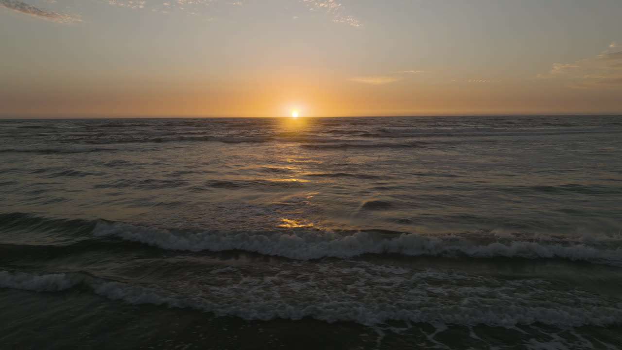 Aerial panoramic trucking pan over waves crashing and rolling to shore as ball of light dips below horizon