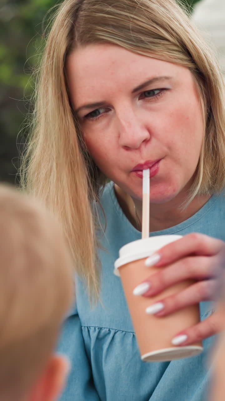 una mujer bebe chocolate de una pajita mientras está sentada al aire libre, mirando enfocada, la vista trasera de las cabezas de dos niños es visible mientras interactúan cerca