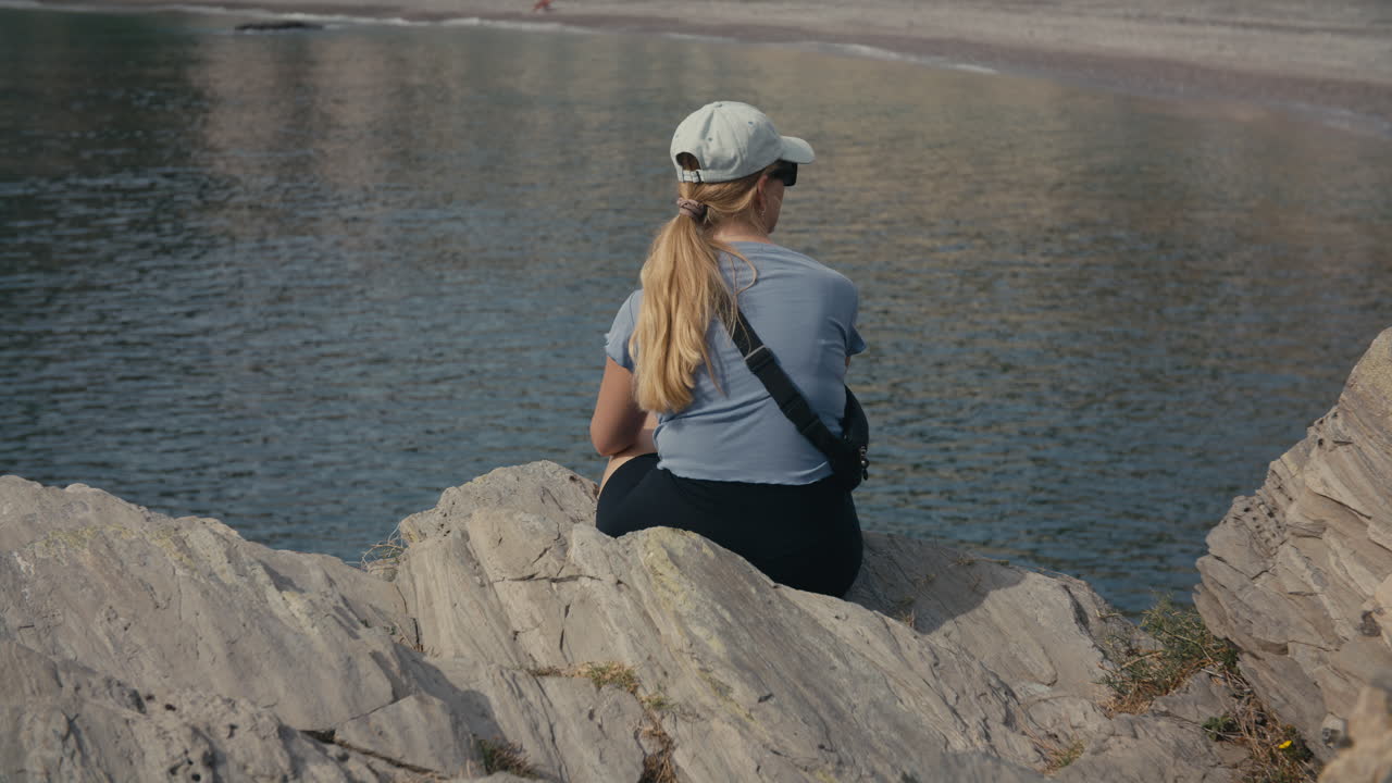 Medium close-up from behind of a woman with blond hair and cap, sitting on coastal rocks, looking out over peaceful sea and shoreline