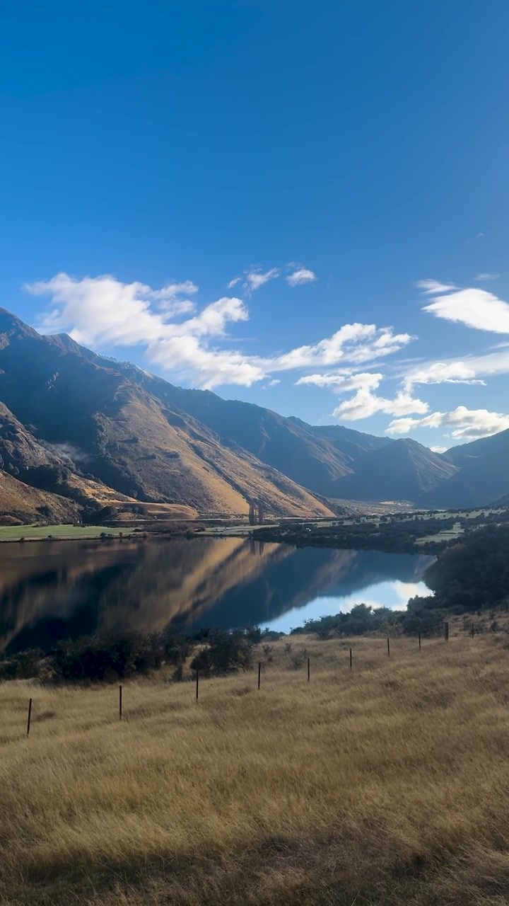 Die Kamera schwenkt über ein grasbewachsenes Feld, einen spiegelnden See und dramatische Berge bei hellem Tageslicht.