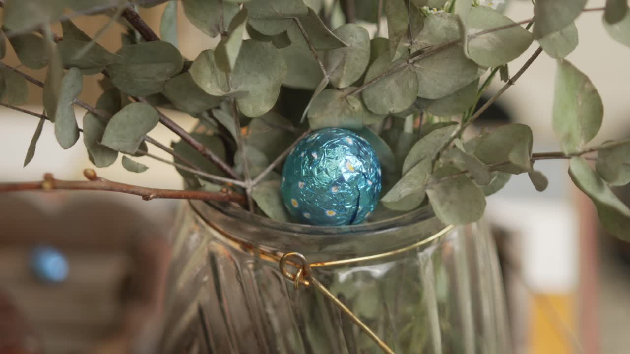 Foil-wrapped blue chocolate Easter egg nestles in eucalyptus bouquet within clear glass vase, creating charming springtime decoration. Soft colors and natural foliage add touch of rustic elegance