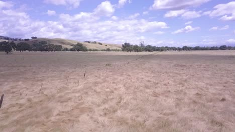 Slow-moving-low-flying-drone-shot-of-rustic-fence-in-a-large-meadow-or-plains-in-golden-California,-flies-along-fence-showing-wide-open-field,-turns-left-to-reveal-stunning-hills-and-cloudy-blue-sky