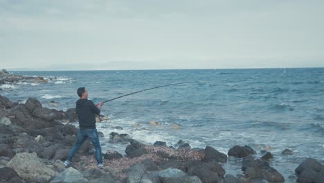 man in difficulty reeling in fishing rod along windy rocky shoreline wide shot