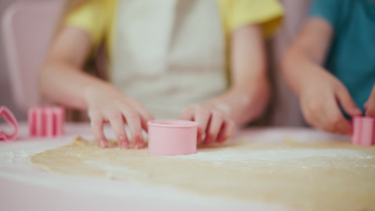 Nahaufnahme von Kindern, die mit einem rosa Plastikausstecher Formen aus flachem Teig stechen. Ihre kleinen, mehlbestäubten Hände arbeiten vorsichtig auf einem hellen Tisch in einer hellen Küche während des Backens.