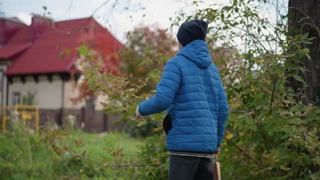 side view of kid with hands in jacket pocket, holding paper bag, approaching tree, plucking leaves and placing them inside bag, red-roofed building in the background, vibrant greenery surrounding