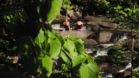 dos mujeres bronceándose en las rocas en medio de un río con las ruinas de un antiguo molino al fondo en el parque gatineau, quebec