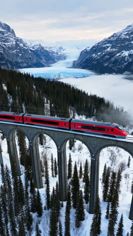 red train crossing a snowy mountain viaduct with a glacier in the background