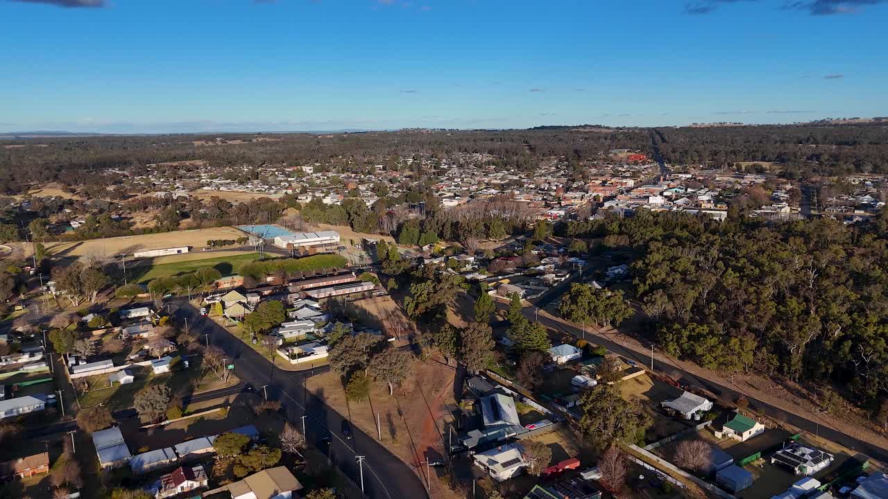 Drone footage glides above a residential area, sports fields, and wooded hills in a rural Australian town under clear daylight, revealing urban layout and natural surroundings