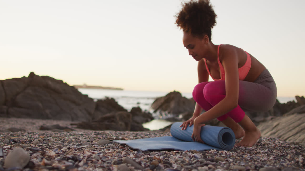 African american woman rolling her yoga mat on the rocks near the sea during sunset
