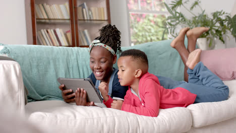 Glückliche-Afroamerikanische-Schwester-Und-Bruder,-Die-Auf-Dem-Sofa-Liegen-Und-Zu-Hause-Ein-Tablet-Benutzen,-Zeitlupe