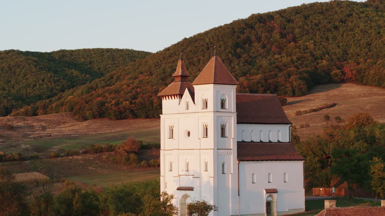 Aerial shot of a white church in Herina, Romania, with a tower, amidst green hills and a winding path