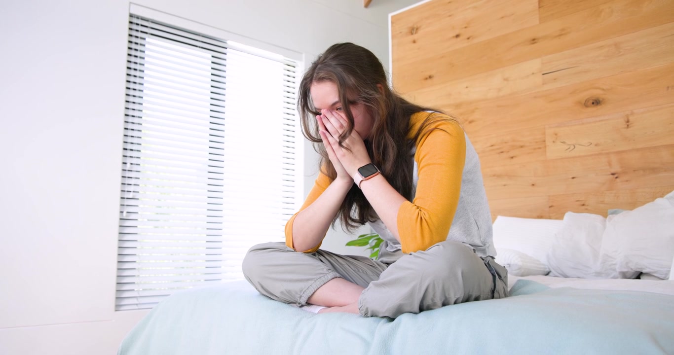 Donna seduta sul letto, lo stress le fa premere le tempie, con le mani giunte, coprendosi il viso con la mano che porta l'orologio.