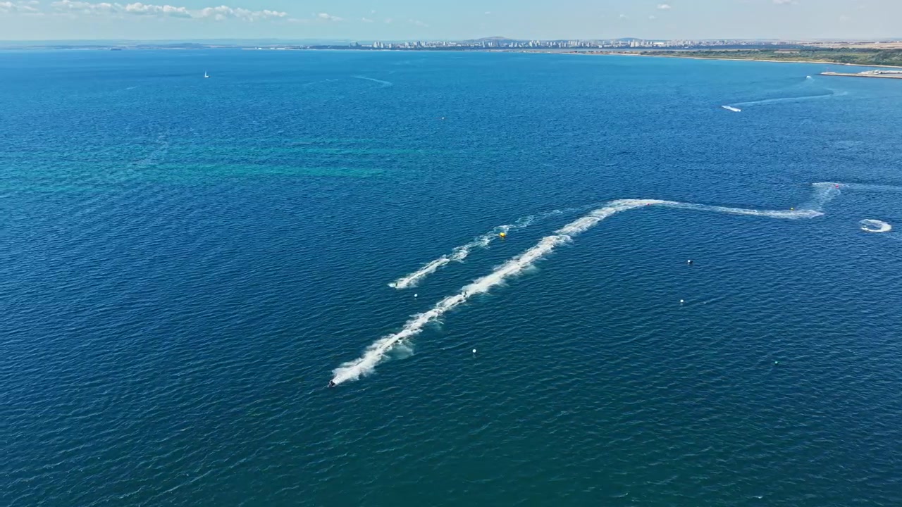 Una gara di moto d'acqua si svolge su un mare blu con cielo limpido e lo skyline di una città in lontananza, in una giornata di sole.