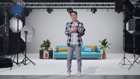 cuerpo lleno de fotógrafo asiático sosteniendo una cámara en sus manos y sonriendo siendo feliz mientras estaba de pie en el estudio
