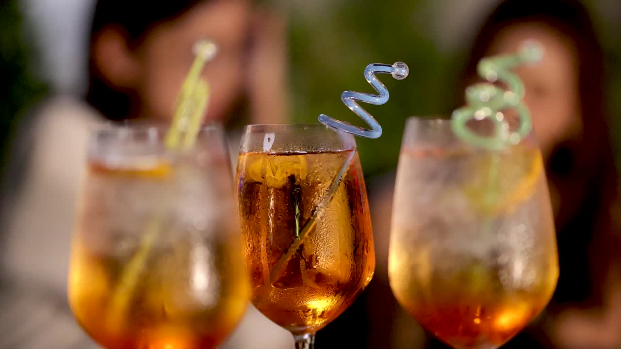 Two friends clink vibrant cocktails with spiral straws, soft focus background, golden hour lighting