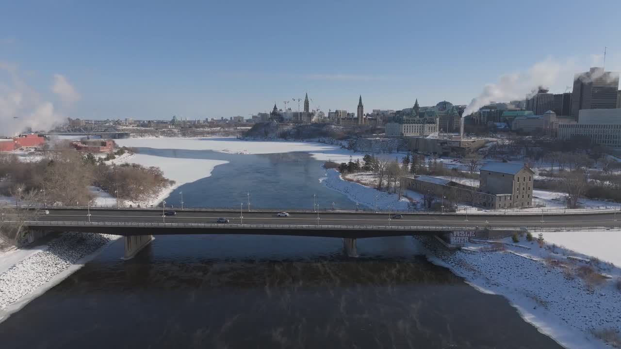 Rekaman drone Jembatan Portage yang melintasi Sungai Ottawa yang membeku dengan Parliament Hill di latar belakang pada hari musim dingin yang cerah dengan kabut yang naik
