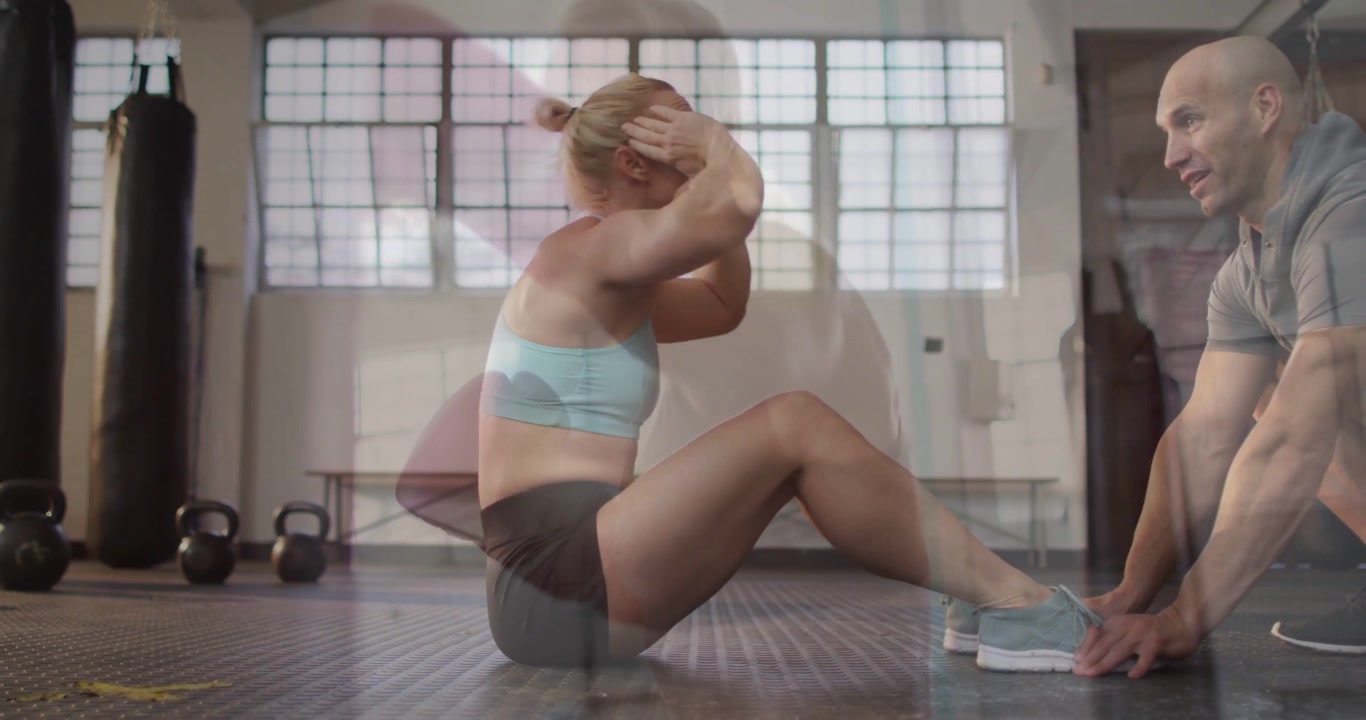 Donna che fa crunch in reggiseno sportivo azzurro e pantaloncini scuri, flettendo il busto in palestra, con l'allenatore che le tiene i piedi.