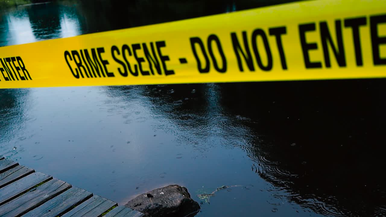 Close up video of a yellow colored Crime Scene Do Not Enter police caution ribbon tape pulled in front of a dark blue lake that is reflecting trees during a rainy evening. Wooden deck at the bottom