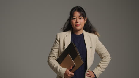 studio portrait of female teacher or businesswoman standing against grey background with folder under her arm 1