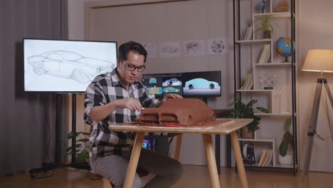 hombre asiático diseñador de automóviles sonriendo y mostrando brazos cruzados gesto a la cámara mientras trabajaba en la escultura de arcilla de coche en el estudio