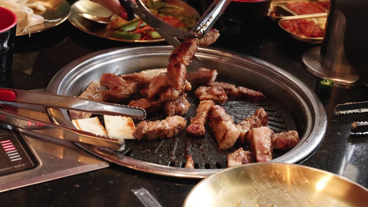 Hands use tongs and scissors to cut pork belly on tabletop grill, warm restaurant lighting