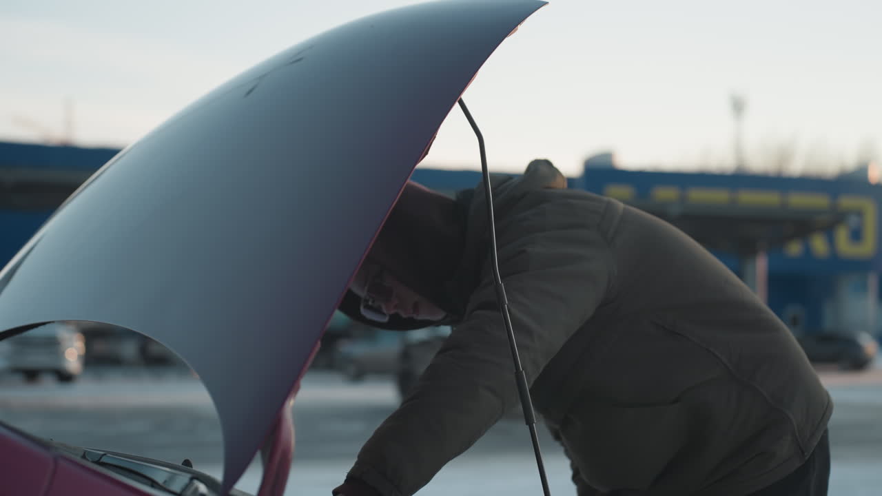Man wearing dark winter jacket leans under raised hood of red car while parked in snowy outdoor lot during cold weather, performing engine inspection or repair with trees and vehicles in background