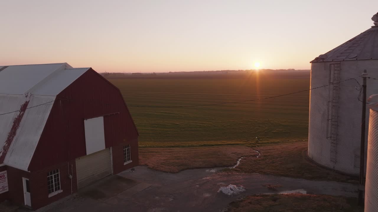 4k drone shot tracking backwards past farmland to reveal a red barn and silos at sunset in the American Midwest