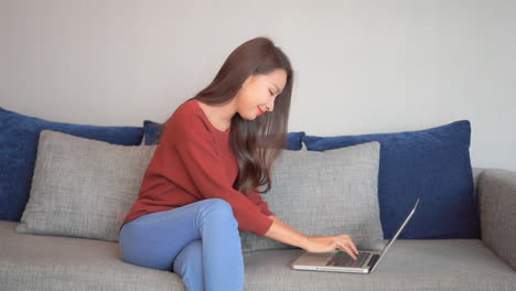 Joven-Asiática-Sonriente-Sentada-En-El-Sofá-Usando-Un-Portátil-Mirando-El-Mensaje-De-Escritura-En-Pantalla,-Señora-Charlando-En-La-Computadora-Navegando-En-Las-Redes-Sociales-De-Internet-Estudiando-O-Trabajando-En-Línea-En-Casa