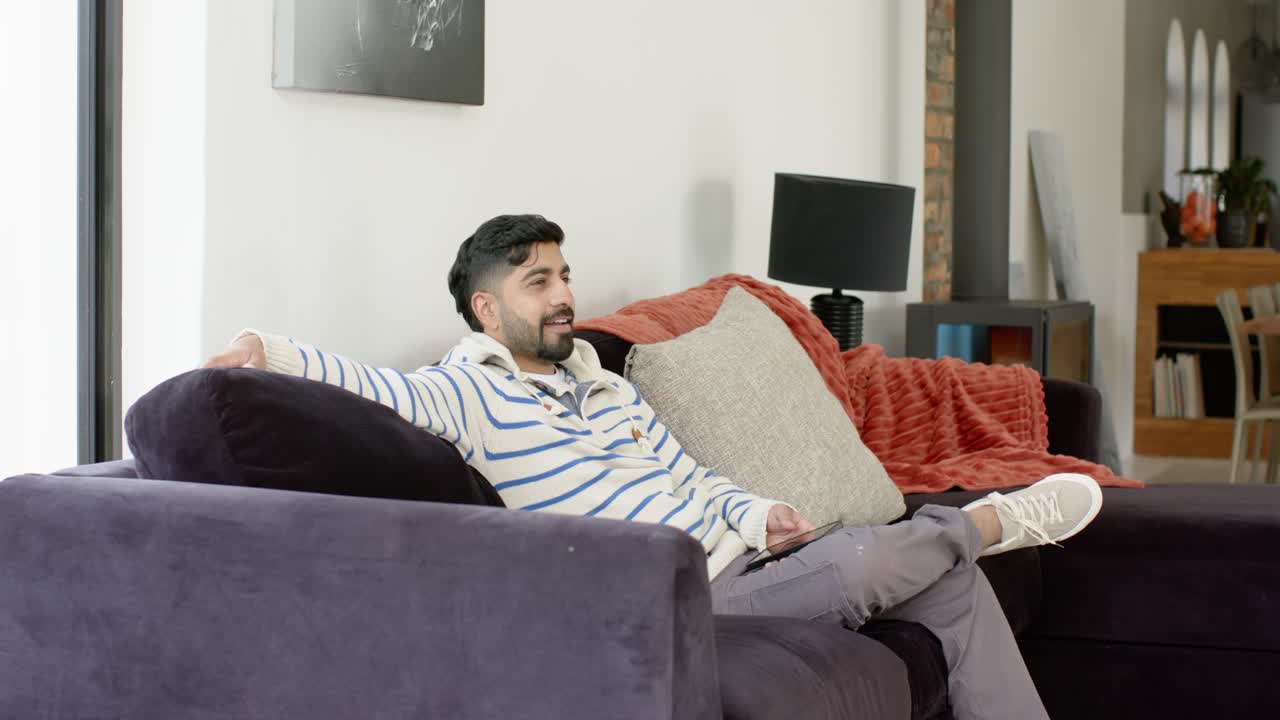 Uomo indiano preme il telecomando, si allunga sul divano in soggiorno mentre aspetta che il programma si carichi