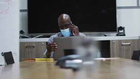 African-american-businessman-in-face-mask-talking-on-smartphone-using-laptop-in-conference-room