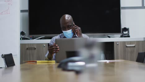 hombre de negocios afroamericano con máscara facial hablando por teléfono inteligente usando una computadora portátil en la sala de conferencias