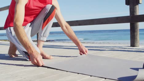 video of happy caucasian man with dreadlocks laying down yoga mat on beach promenade in sun