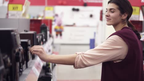 Young-attractive-girl-smiling-and-dancing-while-looking-for-audio-system-and-choosing-speaker-from-a-row-of-variety-in-an-electronics-store.-Bying-household-electronics.