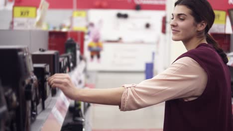 young attractive girl smiling and dancing while looking for audio system and choosing speaker from a row of variety in an electronics store. bying household electronics.