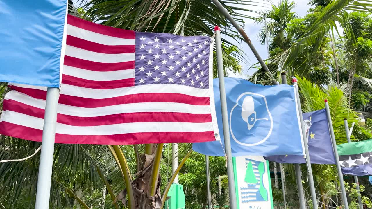 American flag waves prominently outdoors against a backdrop of lush tropical palm trees and foliage