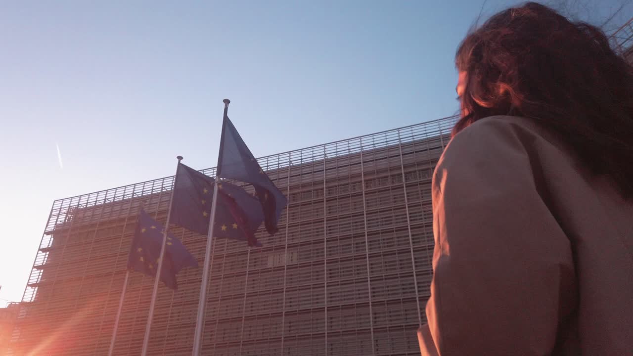 Woman walks under EU flag at dusk. Soft sunlight hits European Commission façade