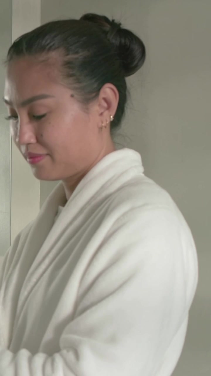 Vertical video: Pressing pump making woman rubbing lotion for skincare at sink, with robe, cabinet