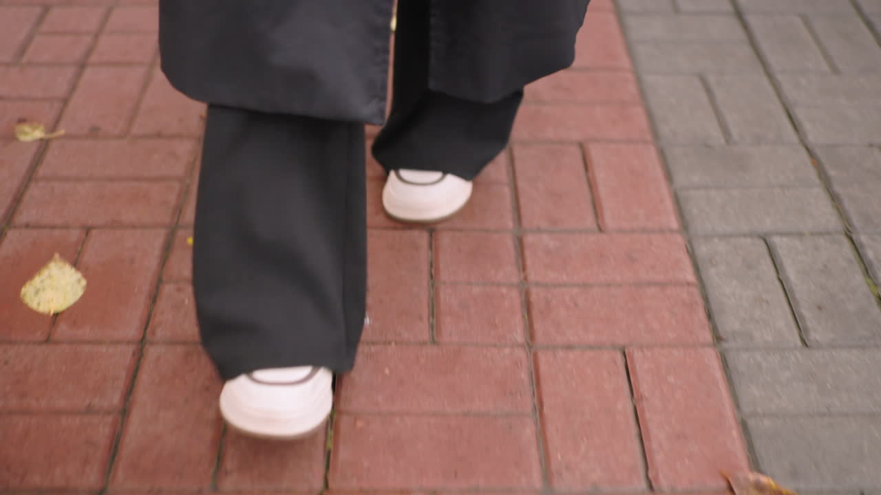 Close-up of woman walking on brick sidewalk wearing black pants and white sneakers, fallen autumn leaves scattered on ground, capturing step movement, casual urban lifestyle scene with seasonal atmosphere