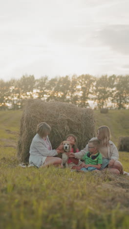 la familia amorosa se reúne en un campo abierto cerca de la bala de heno, acariciando a su perro bajo la puesta de sol dorada, el ambiente cálido y alegre destaca los fuertes lazos, la felicidad y el paisaje rural pacífico