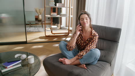 happy young woman sits in modern living room and talks on phone, wide