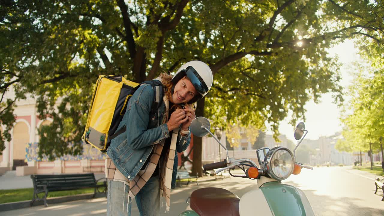 Courier guy with curly hair in a denim shirt puts on a white motorcycle helmet and looks out the side window of his moped along with a big yellow one in a sunny city park in summer