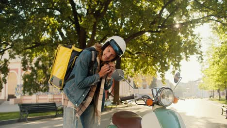 mensajero con el cabello rizado en una camisa de vaqueros se pone un casco de motocicleta blanco y mira por la ventana lateral de su motocicleta junto con un grande amarillo en un parque de la ciudad soleado en verano