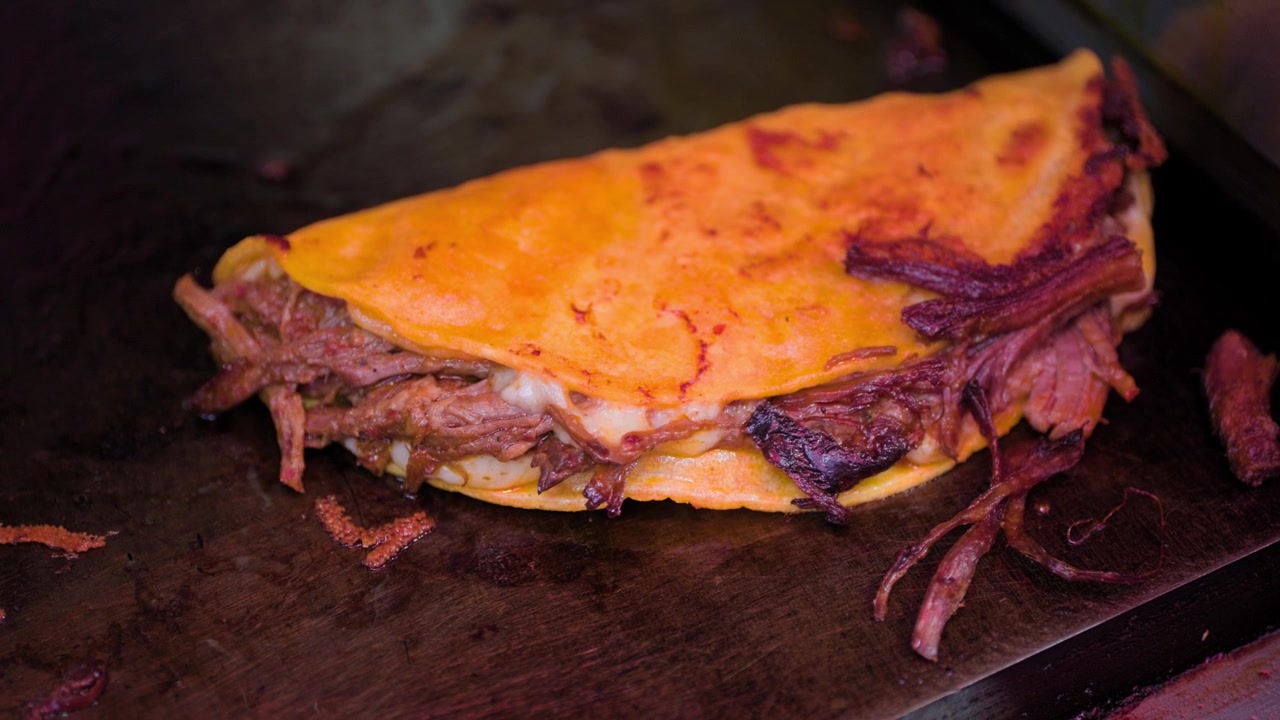 Close-up de um saboroso taco quesabirria sendo preparado em uma chapa quente com queijo derretido e uma tortilha vermelha crocante.