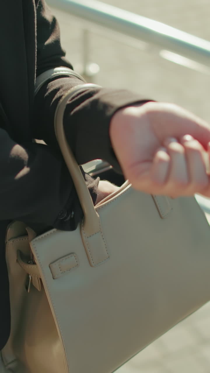 Close up of lady searching handbag outdoors, hand inside bag retrieving item, walking near metal railings, dressed in professional attire with sunlight casting shadows on pavement