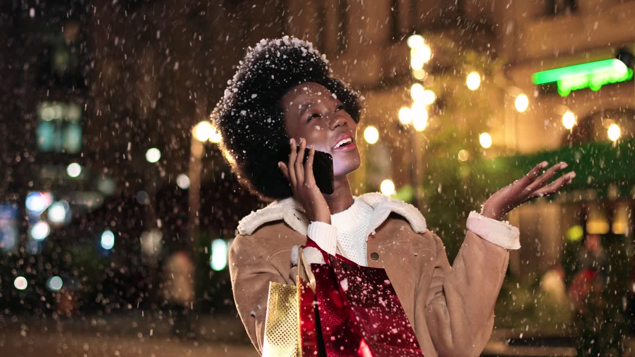 Close-up view of african american woman holding shopping bag and talking on smartphone on the street while it‚Äôs snowing in Christmas