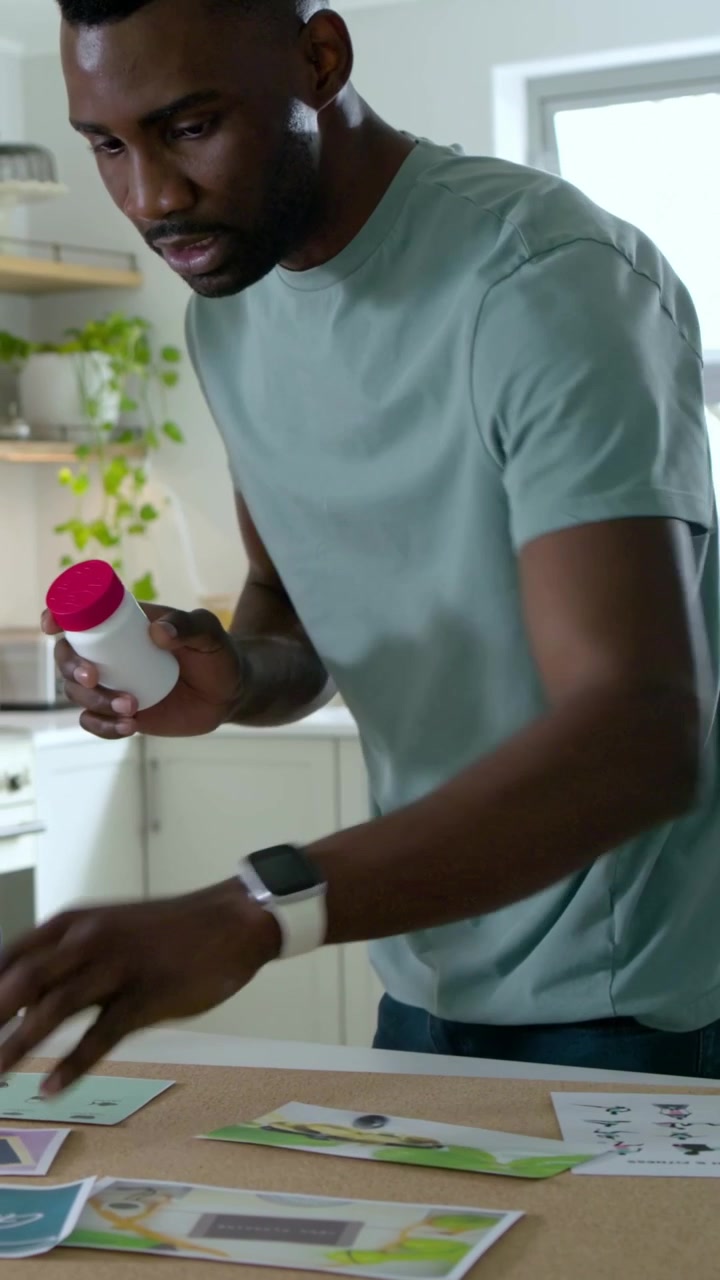 Vertical video: Leaning man in mint tee rotating pink bottle and organizing papers in kitchen