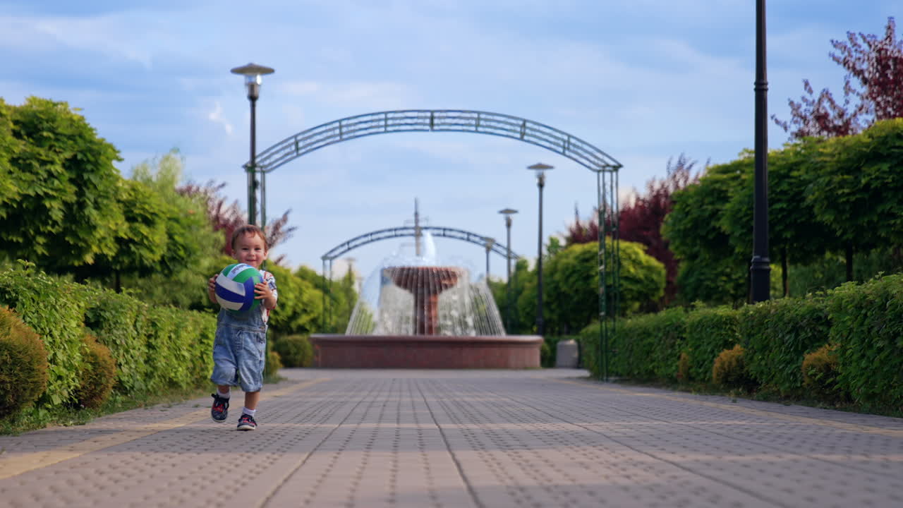 Healthy happy Caucasian baby boy runs by the paved road in the park. Kid approaches the camera and throws a ball.