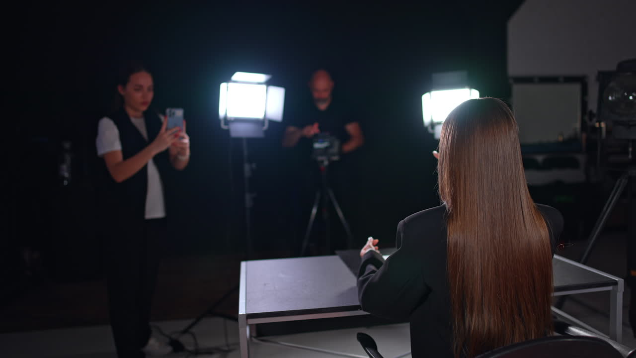 Brunette with long hair sits her back to camera facing soffits. Girl at backdrop taking video on her phone and cameraman uses professional camera. Studio blog footage.