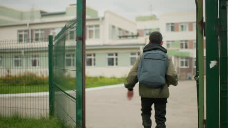 back view of kid wearing backpack running through school gate towards classroom building with focus on determined movement showing energy and purpose in daily routine of student life