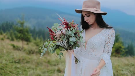 sad girl with flowers in mountains background. young bride in white wedding dress and pink hat holding flowers against distant mountains.